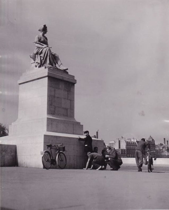 DOISNEAU (Robert). | Des passants s'abritent derrière l'une des statues du pont du Carrousel. Photographie originale en tirage d'époque.